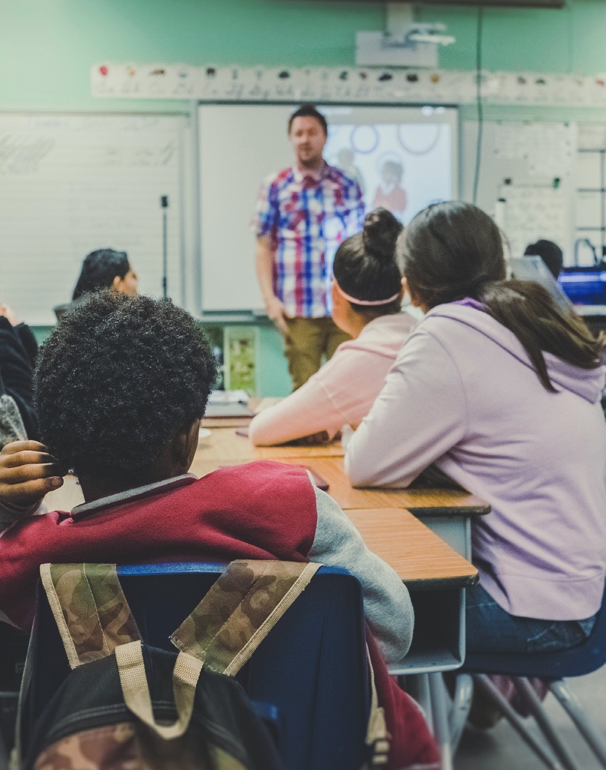 Students seated at desks in a classroom, facing an adult standing near the front